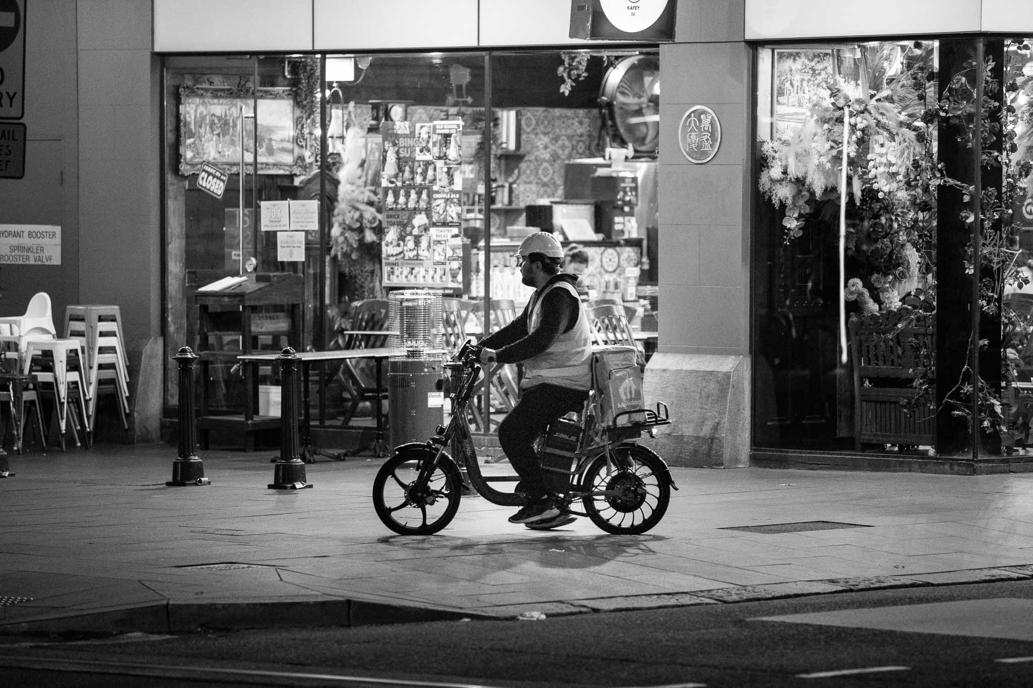 A guy on a delivery ebike dressed in high viz and a hard hat, cycling in front of a closed restaurant. Unused chairs are visible stacked up at edge of frame.