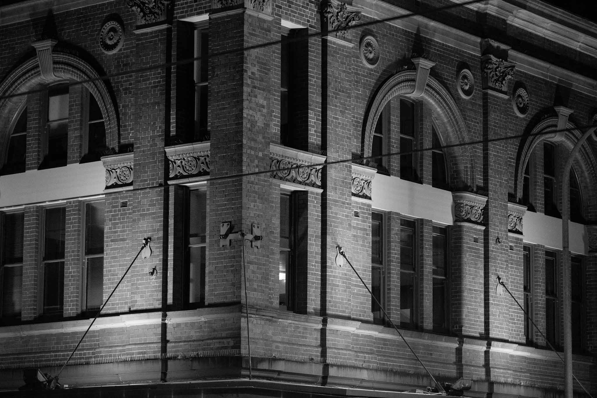 A corner-on view, almost flat, of a brick commercial building in Haymarket. The building has stays connecting the brickwork to the verandah, and also has two-storey high arched windows set into the brickwork. Overhead cables from the tram are out of focus in the foreground crossing the image.