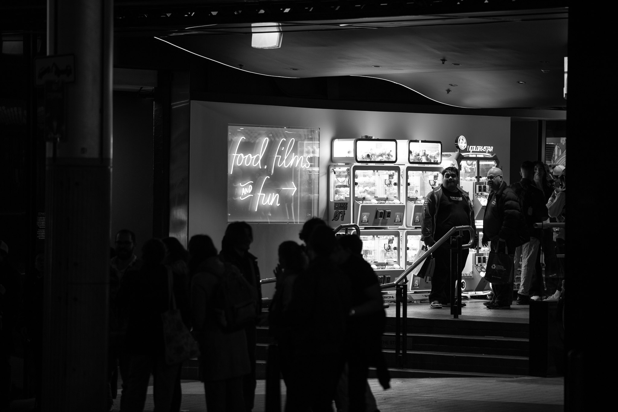 A crowd of a half dozen is in silhouette in front of a few steps, leading up to a wall that has a few miniature Claw Machine games mounted to it. A neon sign reads "food, films and fun", with a couple of guys in front of the claw machines clearly not having food, fun or watching films.