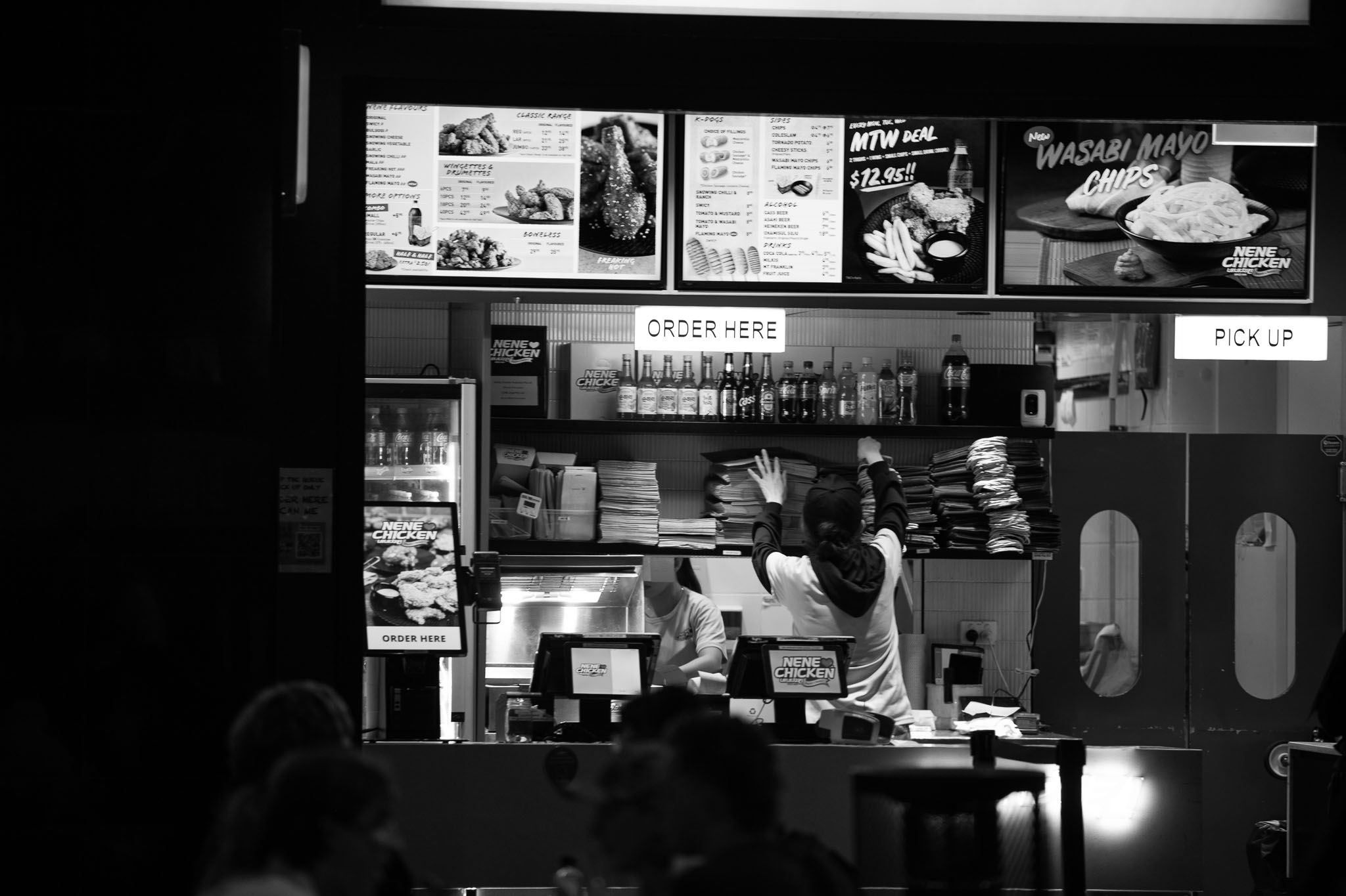 The storefront of a fried chicken kiosk, with a worker on the front register facing away from the camera, reaching up for something. In the background, the faceless body of the kitchen worker can be seen behind the counter, with the tops of a few heads in the foreground, out of focus and in silhouette.