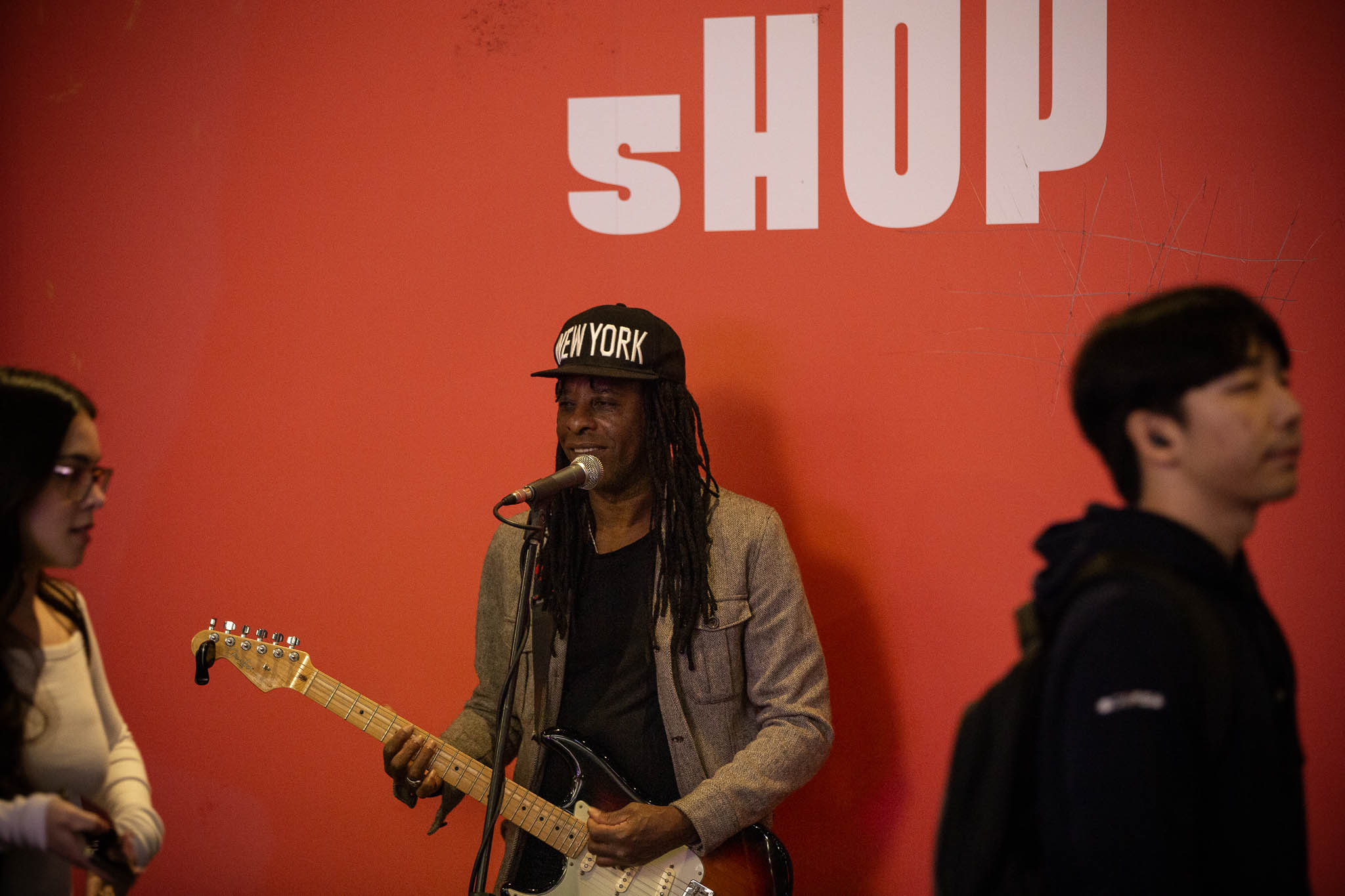 A guitarist is standing in front of a red-coloured wall, with "SHOP" above his head in white, sans serif, heavy text. He's playing an electric 6-string guitar with a white and red body, wearing a grey jacket, black shirt and black cap that says "NEW YORK".
A woman is about to pass him from left of frame, with long dark hair, wearing a white top and carrying a phone. A man has already passed him to the right, out of focus and wearing a dark top.
Neither are taking much notice of the performance.