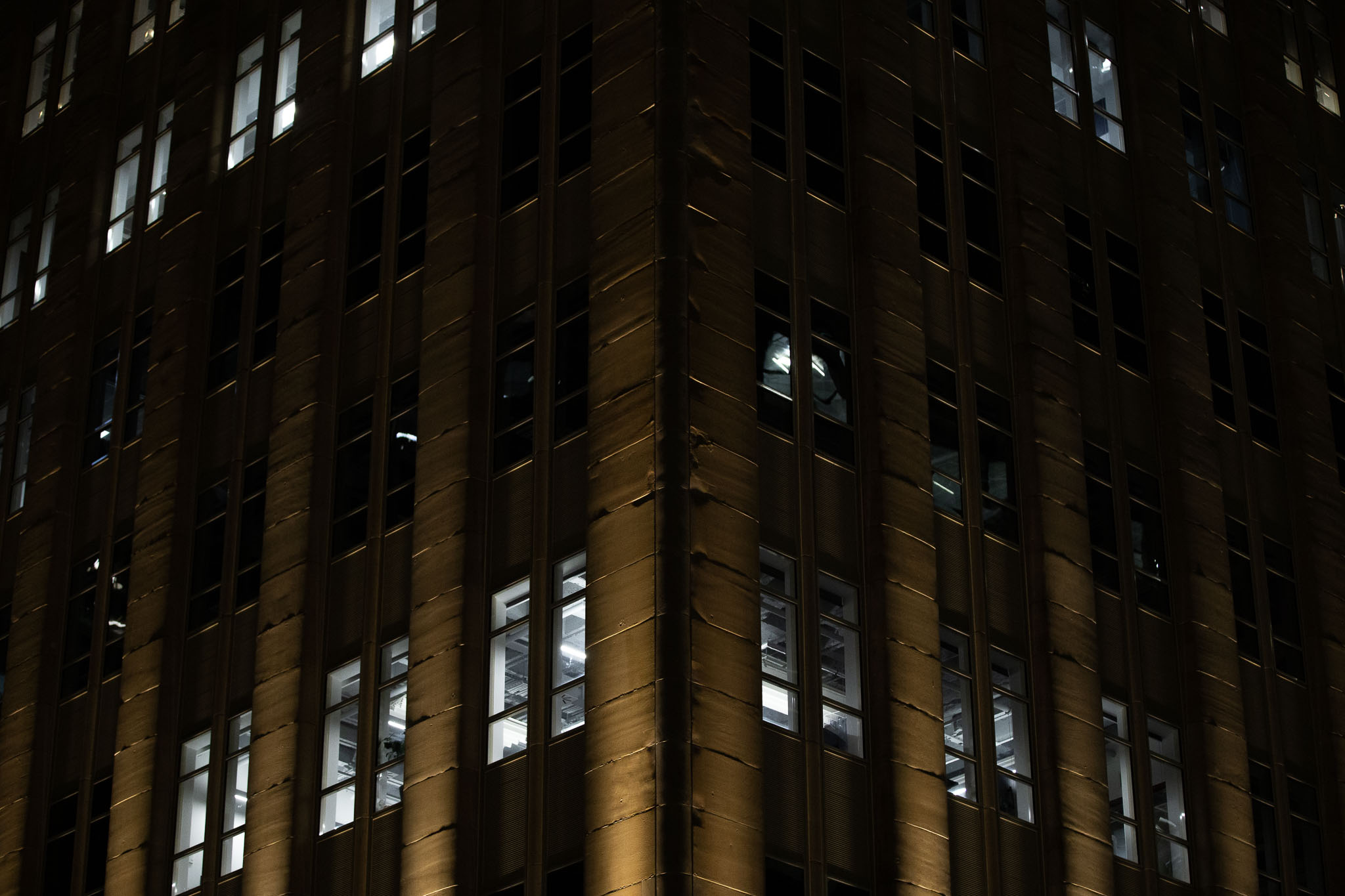 An image of 4 levels of the MLC Building on Martin Place, Sydney, taken from a 45º angle on the corner of the building. Sandstone columns are vertically lit from below with a warm, yellowy light, with a couple of floors showing stripped ceilings and harsh interior light from fluorescent lamps.