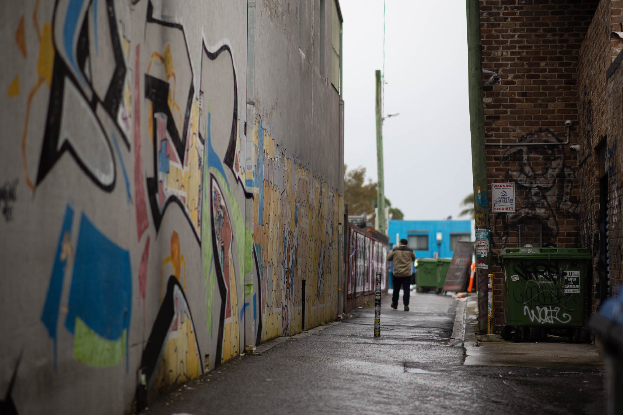 A scene of a pedestrian laneway between two buildings, one wall consisting of a prefab concrete slab, covered in yellow and orange graffiti, with the occasional hint of green, blue and red segments. A man is walking away from the camera in the distance, towards an extension of the laneway beyond the buildings. In the foreground is a single pole in the middle of the laneway, about waist height, and a green dumpster nudged in the corner of the opposite red-brick-clad building in an L-shaped nook. Two security cameras with rectilinear cable conduit are mounted on the brick wall, watching the laneway.