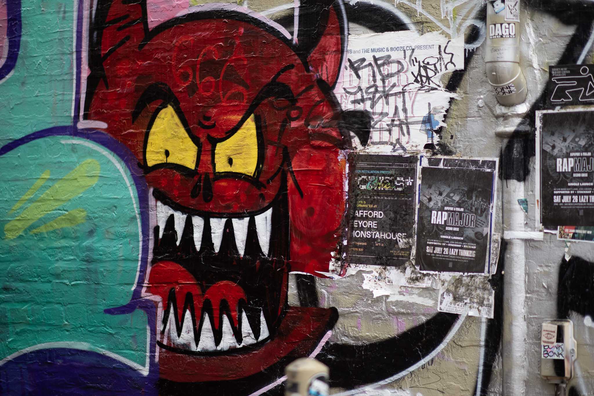 A scene of a laneway wall, with a large, spherical red demon illustrated in cartoon style, with large, yellow eyes, a wide-open mouth with jagged white teeth hiding a tongue. The demon has been overpainted by someone's tag in teal, outlined in blue. Various posters have been stuck up on the wall on the other side.