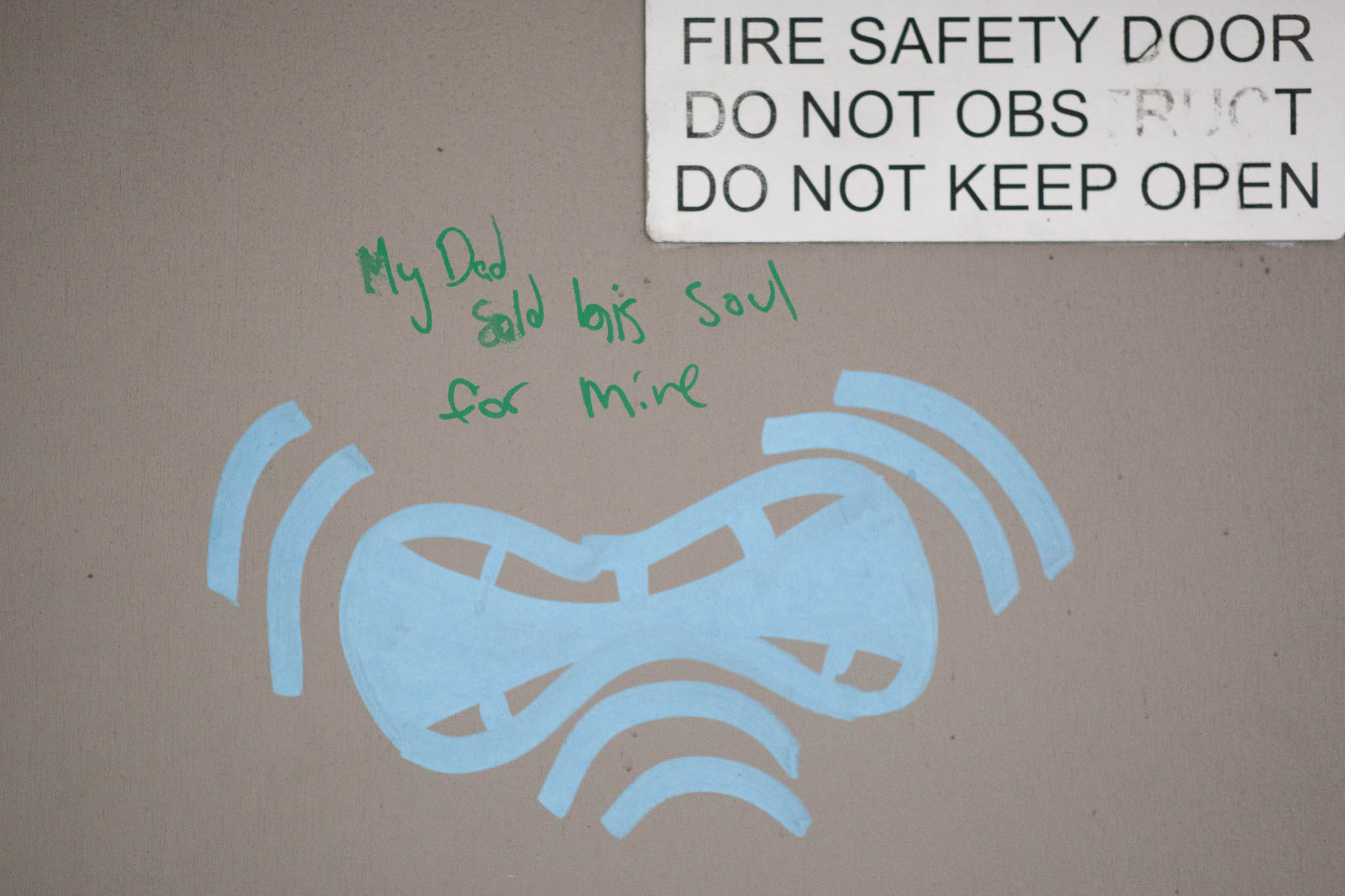 A flat-coloured door painted in beige, with a sign at the top reading 'DO NOT OBSTRUCT. DO NOT KEEP OPEN'. On the door someone has written in green handwriting, 'My Dad sold his soul for mine', and underneath the text, a light blue drawing of figure-of-eight shaped goggles have been drawn inside two motion lines.
