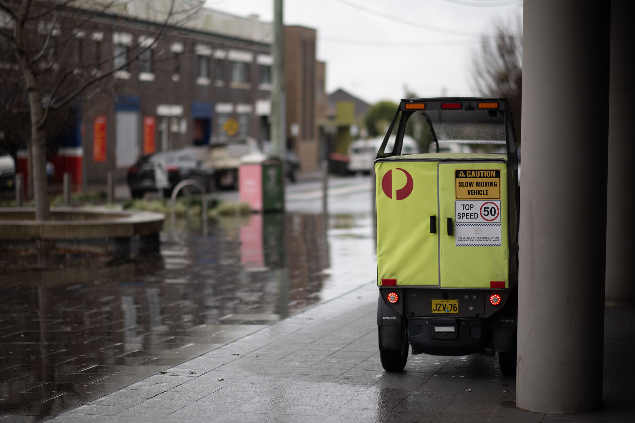 A wet, dreary scene that doesn't show too much active rain, just soaked pavers in a small plaza, the end of a suburban street in the distance with a couple of parked cars. Off to the right is a tiny Australia Post delivery buggy in fluoro yellow, complete with a 'Caution, slow moving vehicle' and 'top speed 50' sign.