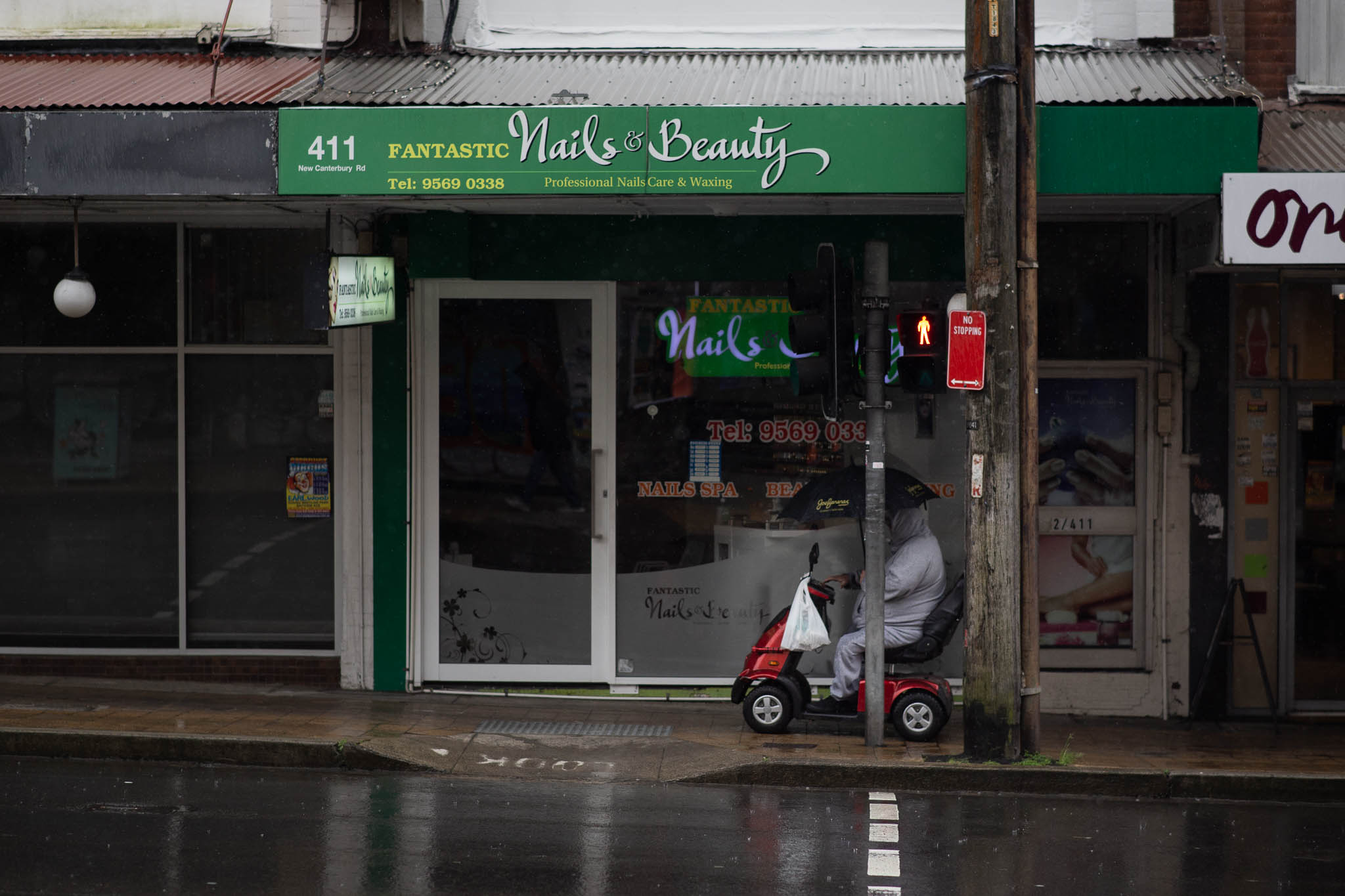 A person is in a mobility scooter, waiting at a pedestrian crossing intersection to cross a major road. Behind them are three storefronts, looking dark and dilapidated, the crossing showing a red light. The person is carrying an unfurled umbrella, and has a shopping bag on the handles of their scooter.