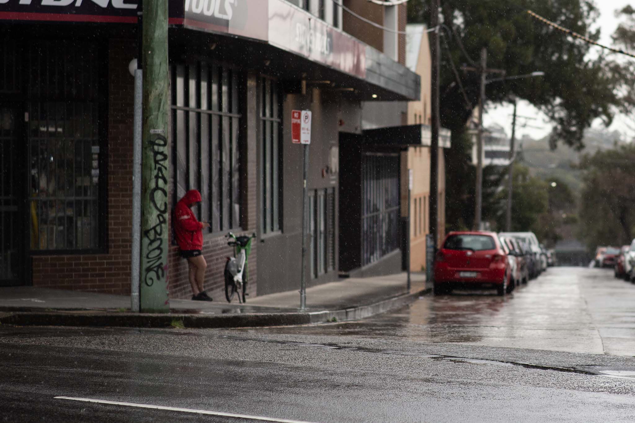 A person in a red puffy jacket is leaning against the side of a brick-clad building, the ground floor housing a retail storefront. The windows are covered with metal bars, and an electricity pole is in the foreground, covered in black graffiti. Next to the building extends a suburban street going downhill, and an overcast scene of blurry, wet houses and parked cars in the background.