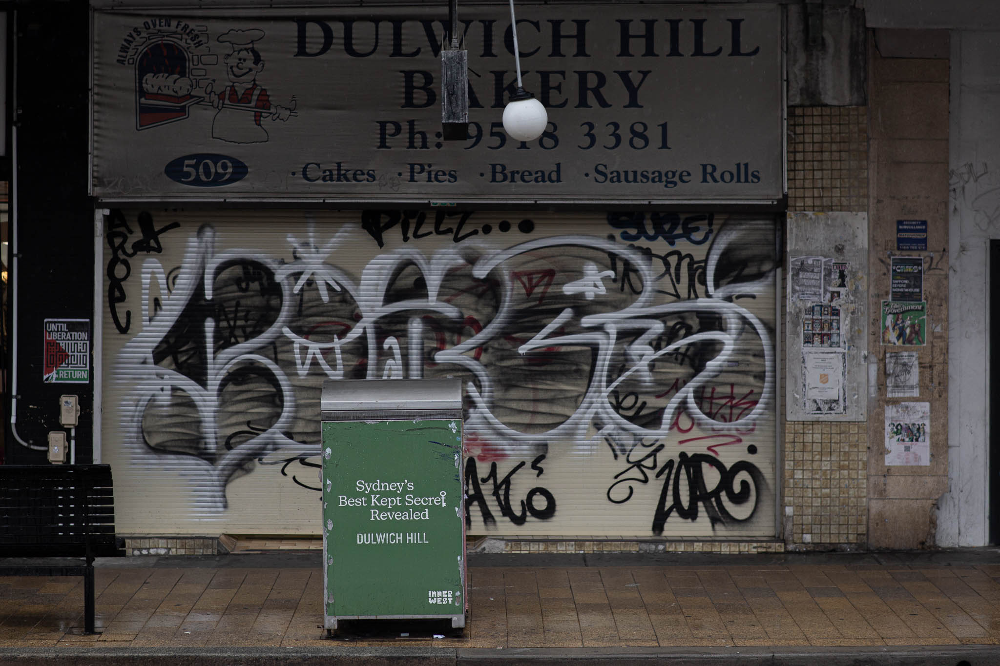A photo of a closed storefront, with metal roller doors shut, and entirely covered with black and silver graffiti. The walls next to the doors are covered with posters, some unreadable, others mentioning Palestinian liberation. Above the storefront is a stretched canvas sign reading 'Bakery, Ph: 9518 3381, Cakes, Pies, Bread, Sausage Rolls'. In the foreground is a metal rubbish bin and the end of a public bench, the bin covered in a council-sponsored wrap reading 'Sydney's Best Kept Secret Revealed, Dulwich Hill'.