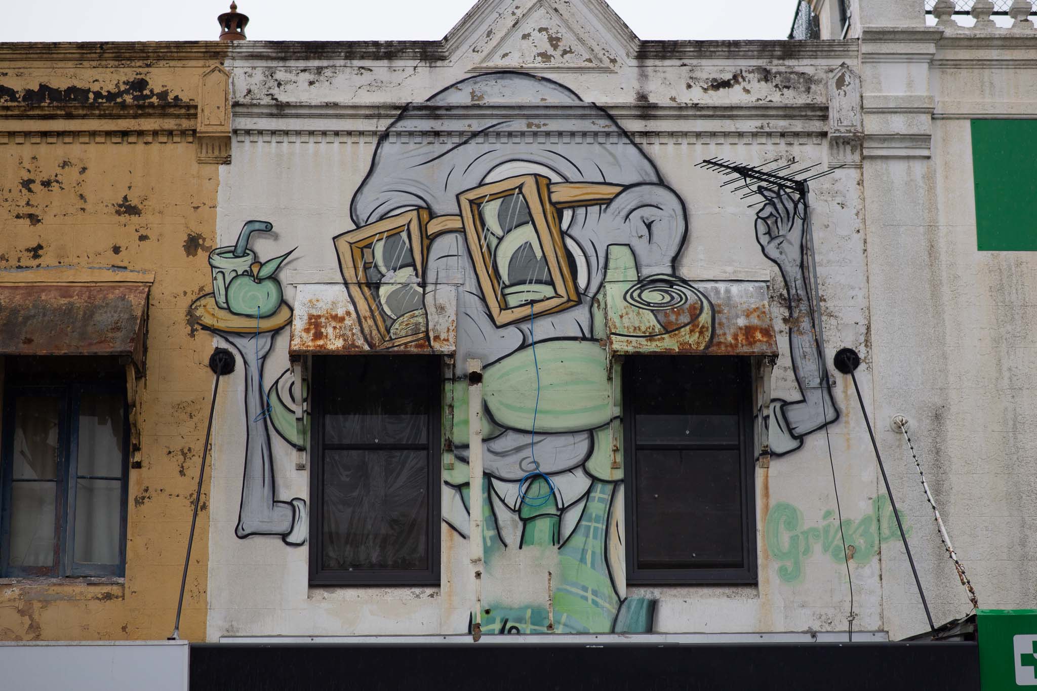 A cartoon-style portrait of a grey-skinned, wrinkled old man is painted across the upper floor of a one-up-one-down against a cream-coloured background, two regular-sized windows framing either side of the portrait. The man has a green, curly moustache, one hand holding a log-periodic antenna, the other holding up a platter with a drink and an apple, his torso wearing a tartan vest, shirt and tie. The building to the left is a strongly-saturated yellow facade.