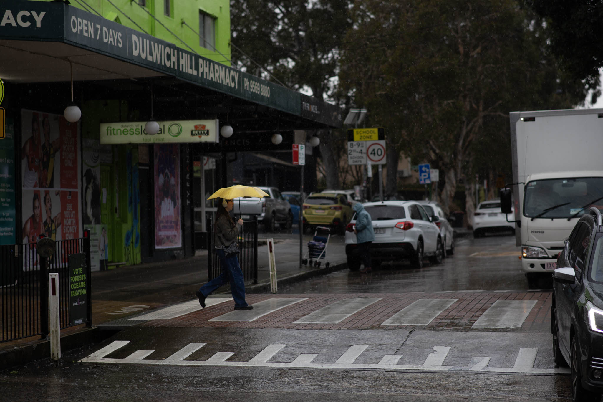 A woman is walking across a zebra crossing, mid-stride, carrying a bright yellow umbrella. Her face and torso is in shadow, but the umbrella stands out from the dark storefront behind her. The left side of the street is clear, but the right-hand side has stopped traffic, a black SUV in front of a commercial truck leaving a narrow gap across the raised pedestrian crossing.